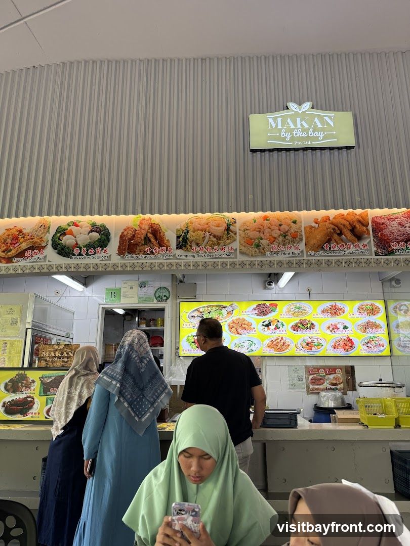 A food stall with a prominent sign reading "MAKAN by the bay Pte. Ltd." above a display of various dishes. Below the sign, a row of food images with descriptions such as "Sweet & Sour Fish", "Broccoli w/ Scallops", "Golden Fragrant Crayfish", "Kampong Thick Bee Hoon Mee", "Shrimp Fried Rice", and "Crispy Prawn Paste Chicken Wing". A menu board with multiple food pictures is visible behind a man ordering. Several people are seen in the foreground, some looking at their phones.