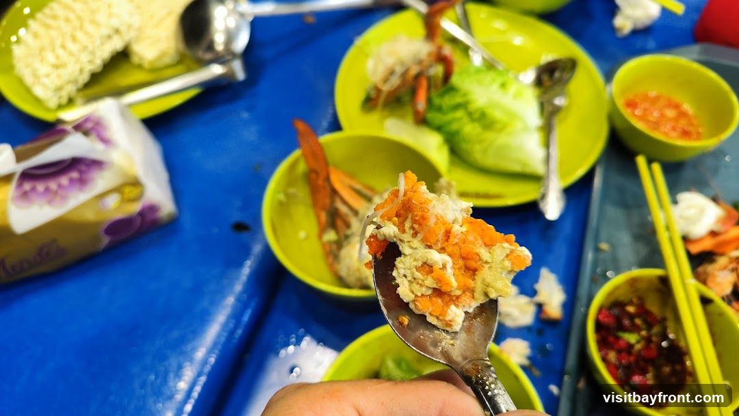 A close-up shot of a seafood meal on a blue table. A spoon holds a portion of crab meat mixed with roe and a creamy sauce. In the background, there are plates with crab legs, lettuce, instant noodles, and small bowls of dipping sauce. A packet of tissues is also visible.
