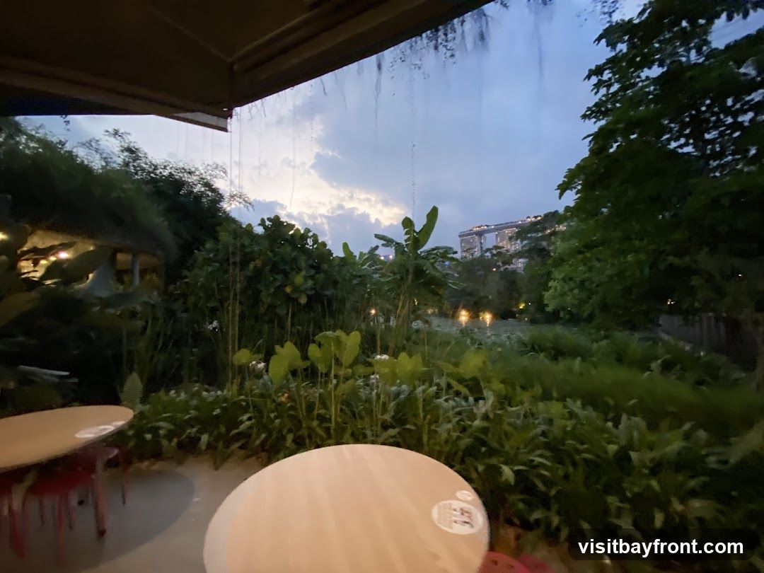 An outdoor seating area with round tables and red chairs is visible in the foreground. Beyond the seating area, lush greenery and trees create a natural ambiance. In the distance, the iconic Marina Bay Sands hotel complex is illuminated against the twilight sky. The scene suggests a relaxed dining or lounging experience with a scenic backdrop.