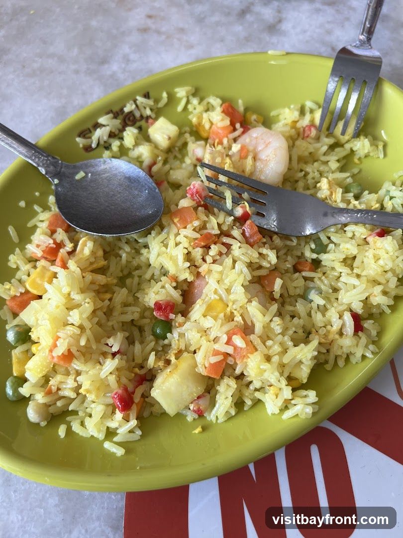 A close-up shot of a plate of shrimp fried rice, with a spoon and two forks resting on it. The rice is mixed with peas, carrots, corn, and pieces of pineapple. A single shrimp is visible in the dish. The plate has a subtle embossed pattern on its rim. A red and white sign with the word "NO" is partially visible at the bottom of the image.