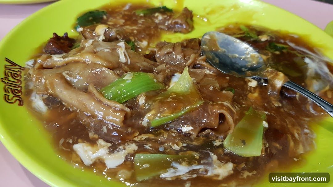 A close-up shot of a plate of satay noodles, a dish featuring wide noodles in a savory brown sauce with pieces of meat and green vegetables. The word "Satay" is visible on the side of the bright green plate.