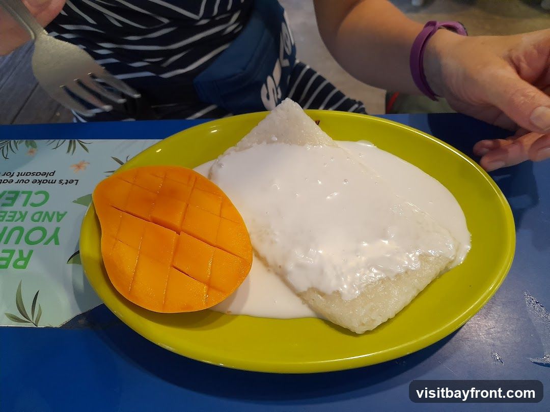 A close-up shot of mango sticky rice served on a bright yellow plate. The dish features a portion of sticky rice topped with a creamy white sauce, accompanied by a perfectly sliced ripe mango. The plate rests on a blue table, with a placemat featuring green text and floral designs visible on the left. A person's hands are visible in the frame, one holding a fork, suggesting the meal is about to be eaten.