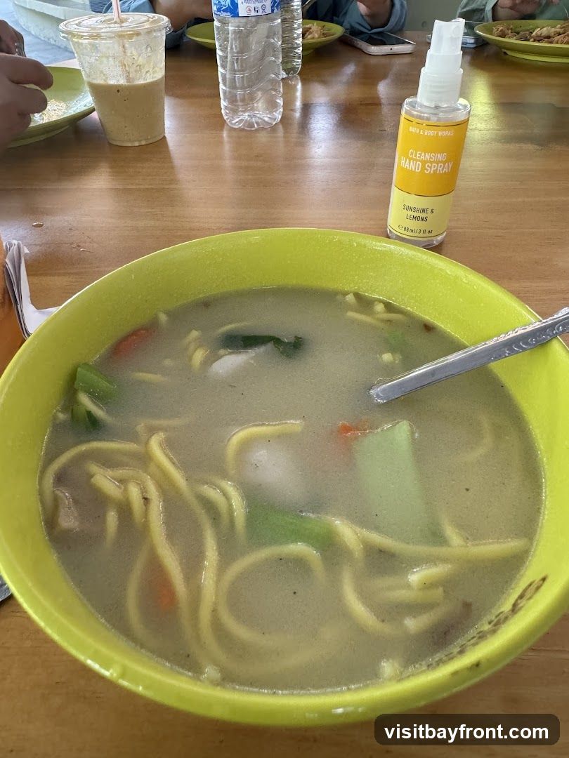 A close-up view of a bowl of soup with noodles and vegetables, placed on a wooden table. A bottle of Bath & Body Works Cleansing Hand Spray is visible to the right of the soup. In the background, there are drinks and plates of food, suggesting a dining setting.