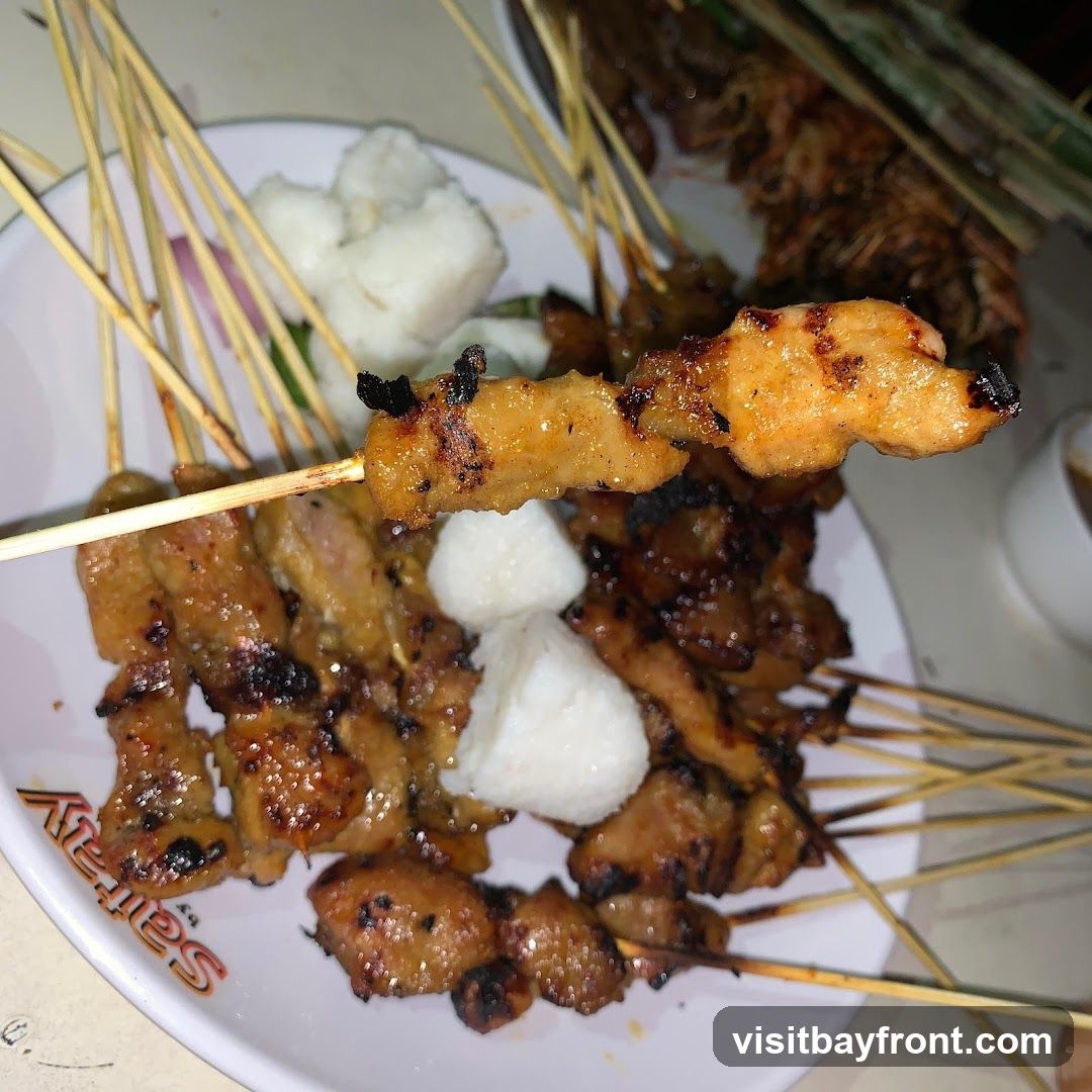 A close-up shot of a plate filled with grilled chicken satay skewers. Some of the skewers are topped with small cubes of white rice cakes. The satay appears to be glazed and slightly charred. In the background, more satay skewers and a small bowl of sauce are visible. The word "Satay" is visible on the rim of the plate.