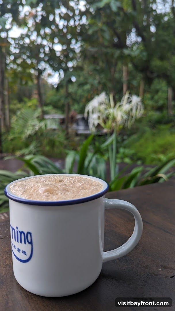 A close-up of a frothy coffee in a white mug with blue trim, set against a blurred background of lush greenery and white flowers. The mug has blue lettering on its side.