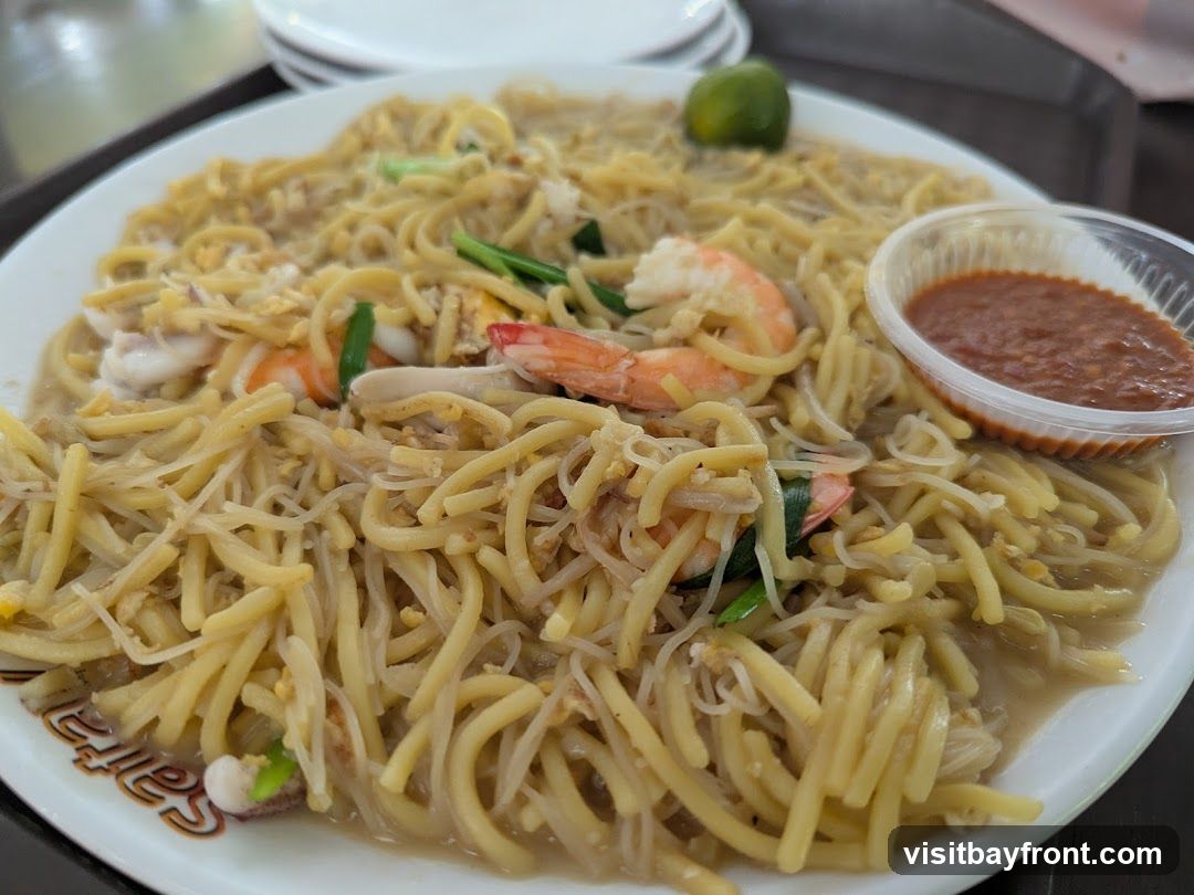 A close-up shot of a plate of noodles with shrimp, squid, and scrambled egg. A small plastic container of sambal sauce is on the side, along with a lime wedge. The plate has text on the rim that is partially visible.