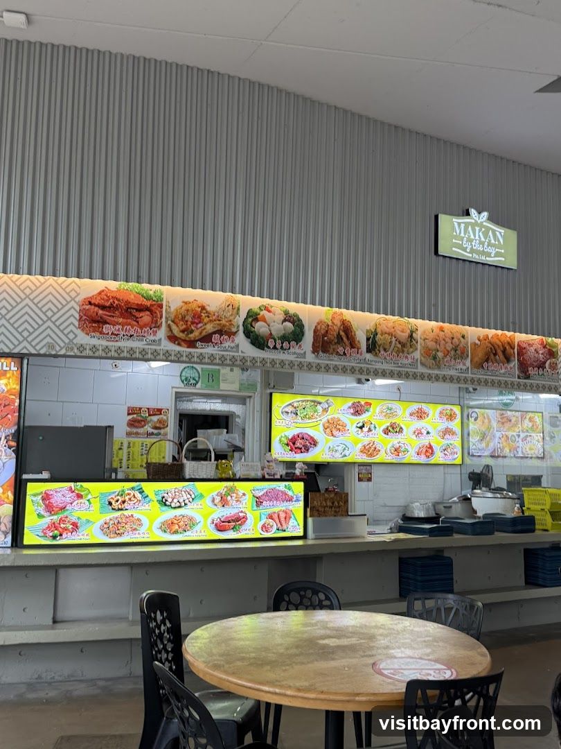 A food stall with multiple illuminated menu boards displaying various dishes. The stall has a sign that reads 'MAKAN by the bay Pte Ltd'. In the foreground, a round wooden table with chairs is visible.