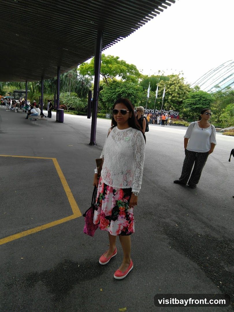 A woman in a white lace top and floral skirt stands in the foreground, looking towards the camera. Behind her, other people are visible, along with lush greenery and the distinctive domes of Gardens by the Bay. The scene appears to be an outdoor walkway with a covered area to the left.