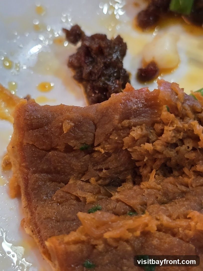 A close-up shot of a braised meat dish on a white plate. The meat appears tender and shredded, with a rich, dark sauce. Small flecks of green herbs are visible throughout the dish. The plate has remnants of sauce and oil, suggesting it's a serving from a meal.