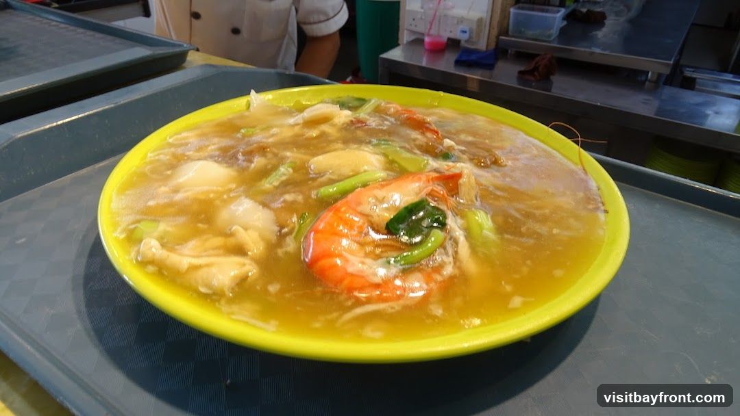 A close-up shot of a bowl of noodles with a thick, savory broth. The dish features large, bright orange shrimp, pieces of white fish or tofu, and green vegetables. The bowl is yellow and sits on a grey tray. In the background, a person in a chef's uniform is visible, along with stainless steel counters and containers.