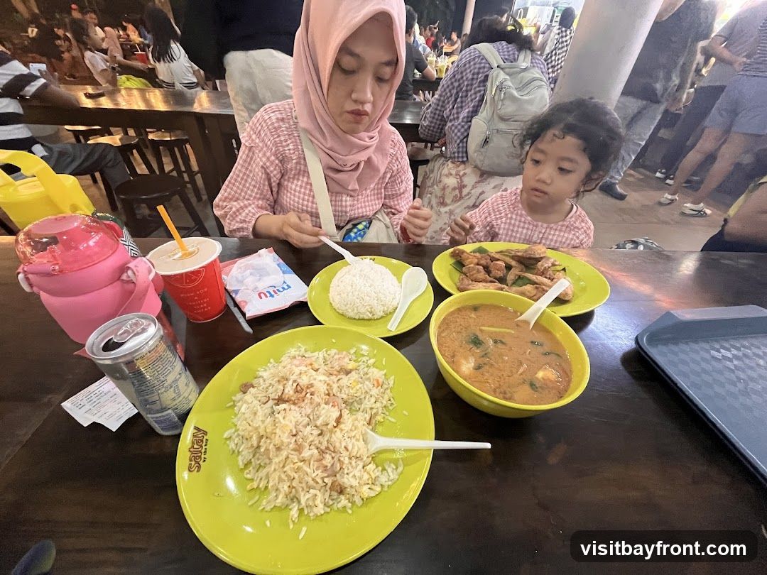 A family is enjoying a meal at an outdoor food stall. In the foreground, a large plate of fried rice is visible, along with a bowl of soup, a plate of fried food, and a bowl of white rice. A child and an adult are seated at the table, with the adult wearing a pink hijab and a pink checkered shirt. Drinks and a can of soda are also on the table. In the background, other patrons are seated at tables, and the atmosphere appears to be lively and casual. The word "Satay" is visible on the plate of fried rice.