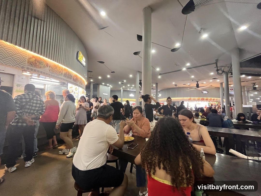 A bustling food court with many people dining and queuing. The food stalls have illuminated signs and displays of food. Tables are occupied by diners, and there is a general atmosphere of activity.