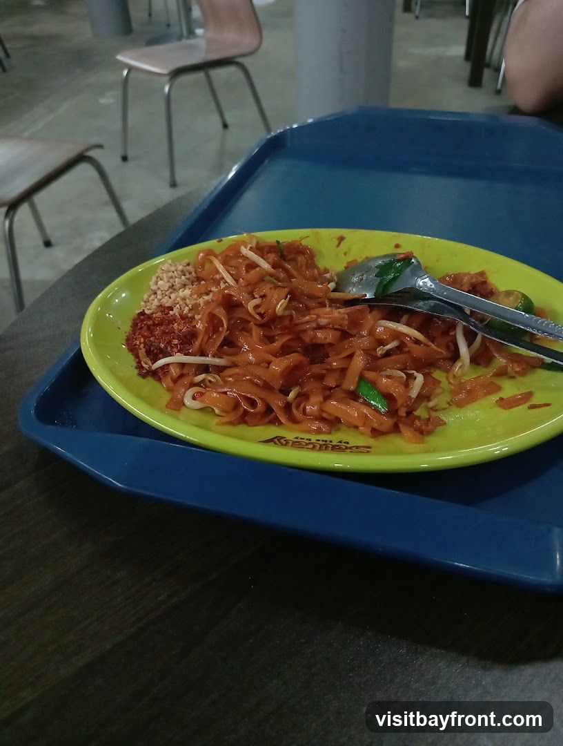 A plate of Pad Thai noodles served on a blue tray. The noodles are mixed with bean sprouts and green onions, and topped with crushed peanuts and chili flakes. A fork and spoon are resting on the plate. In the background, there are chairs and tables, suggesting a dining area. The plate has a logo that reads "Satay by the Bay" and "by the Bay" below it.