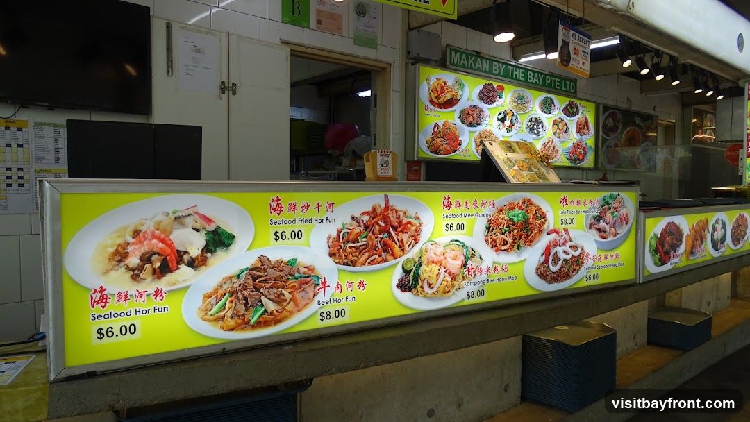 A brightly lit display board showcases various dishes offered at a food stall. The display features images of food items with their names in English and Chinese, along with prices. Some of the visible dishes include Seafood Hor Fun, Beef Hor Fun, Seafood Fried Hor Fun, Seafood Mee Goreng, Kampong Bee Hoon Mee, Lolo Thick Bee Hoon Soup, and Sambal Seafood Fried Rice. The stall's name, "MAKAN BY THE BAY PTE LTD," is visible above the menu.