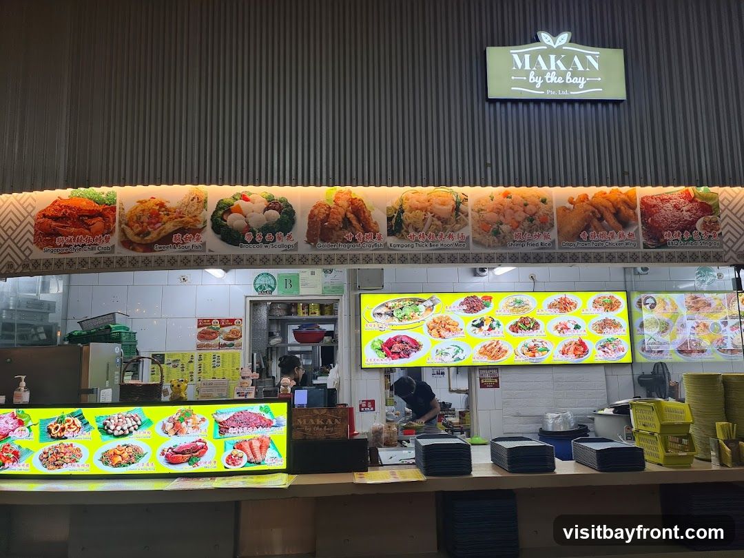 A food stall with a large display of food images and a prominent sign that reads "MAKAN by the Bay Pte. Ltd.". The stall features multiple illuminated menu boards showcasing various dishes, including seafood, fried rice, and chicken wings. A person is visible working behind the counter. Stacks of plates and yellow baskets are also present.