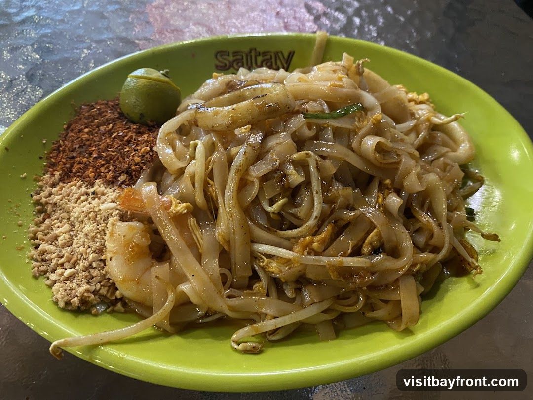 A plate of Pad Thai noodles with shrimp, served with crushed peanuts, chili flakes, and a lime wedge. The dish is presented on a bright green plate with the word "Sataw" visible on the rim.
