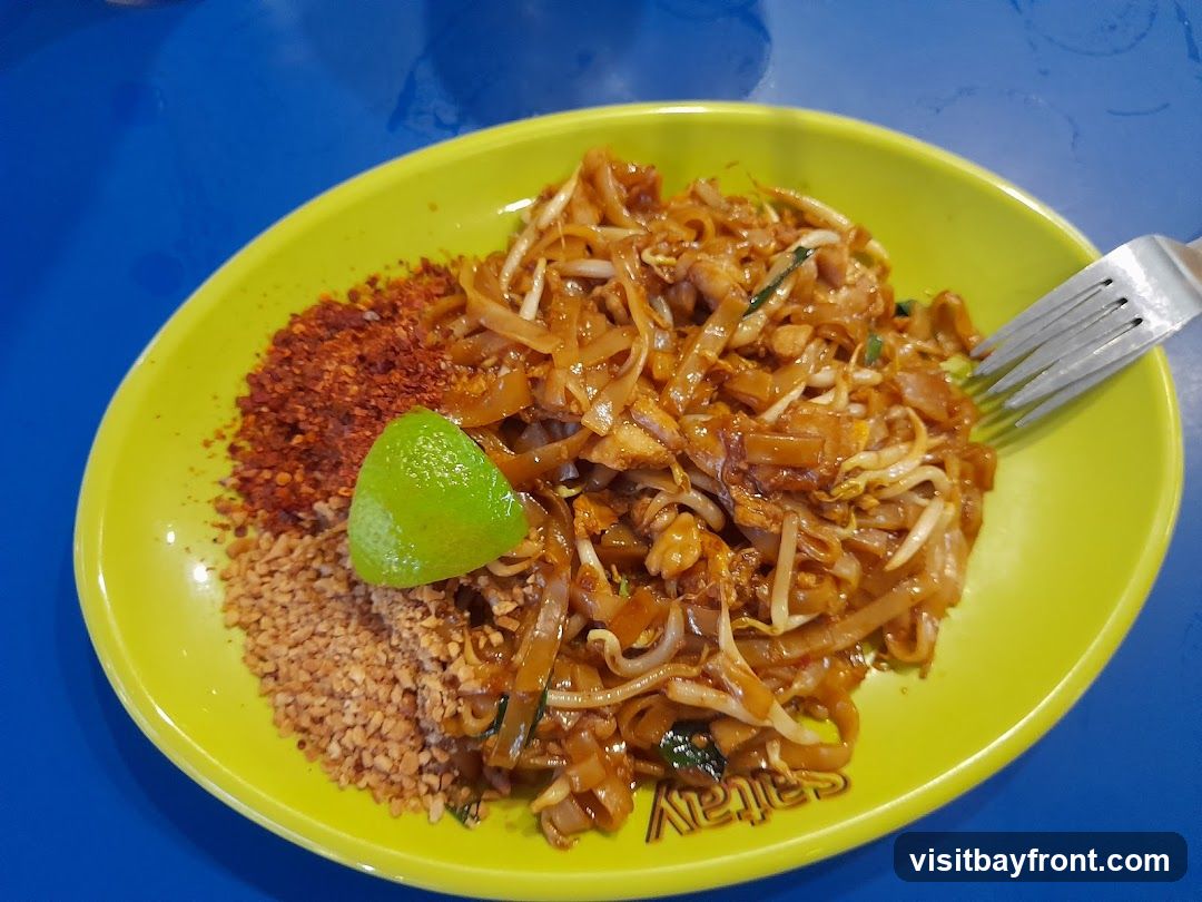 A close-up shot of a plate of Pad Thai noodles served on a bright yellow plate. The noodles are mixed with bean sprouts and what appear to be small pieces of meat or tofu. To the left of the noodles are two piles of condiments: crushed peanuts and red chili flakes. A lime wedge is placed on top of the noodles. A fork is visible on the right side of the plate, partially submerged in the noodles. The plate is set on a vibrant blue surface. The word "Sattay" is visible on the rim of the plate.