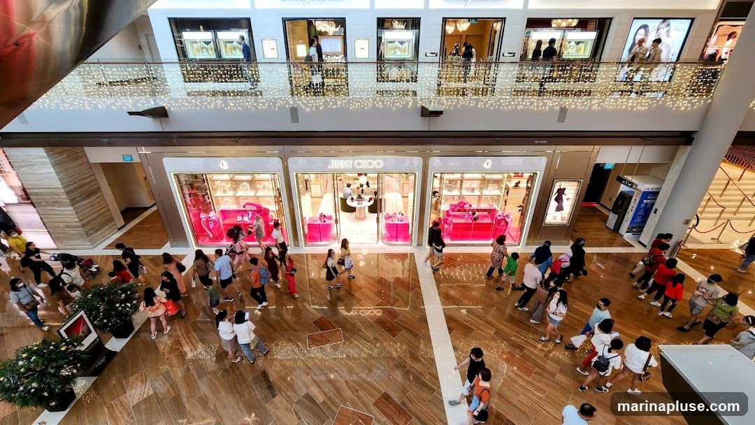 An overhead view of a bustling shopping mall with multiple levels. The foreground features a Jimmy Choo store with its distinctive pink displays. Shoppers are seen walking on the polished wooden floor. Above, other luxury stores like Chopard are visible on the second level, adorned with decorative lights.