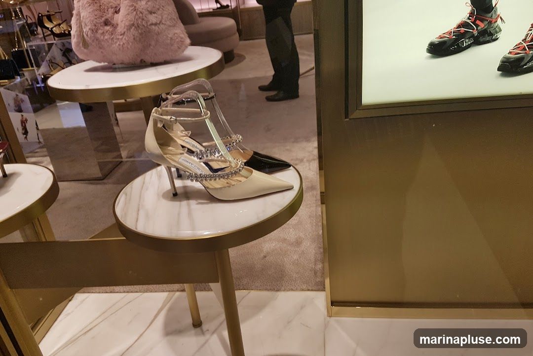 A display of designer high-heeled shoes and a fluffy pink handbag in a luxury retail store. The shoes are showcased on marble-topped tables with gold accents. In the background, a person in dark clothing is visible, and a display of sneakers is seen on a bright white panel.