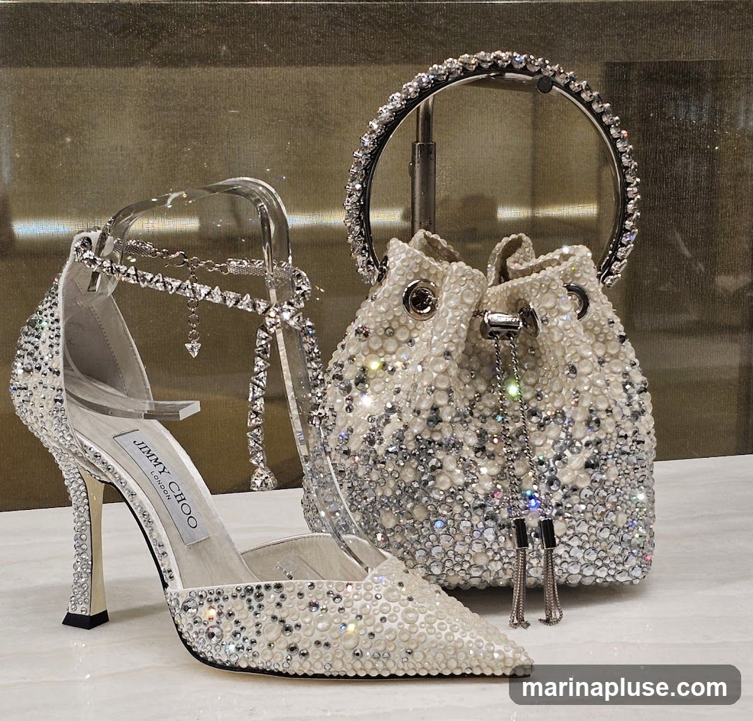 A close-up view of a white stiletto heel shoe and a matching bucket bag, both heavily embellished with rhinestones and pearls. The shoe has a clear ankle strap and a silver heel. The handbag features a circular silver handle and a drawstring closure. The Jimmy Choo London logo is visible on the insole of the shoe. The display is set against a textured gold background.