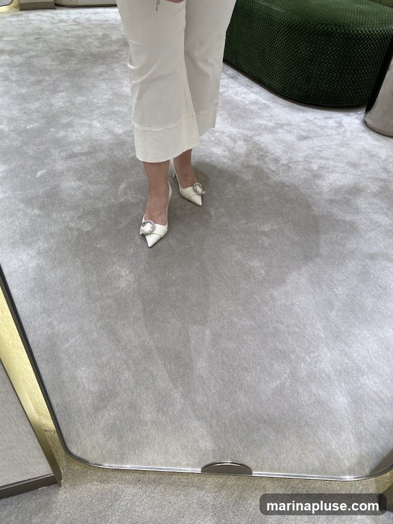 A person wearing white cropped pants and white pointed-toe heels with large crystal embellishments on the front. The shoes are reflected in a mirror on the floor. The background shows a plush grey carpet and a dark green textured ottoman.