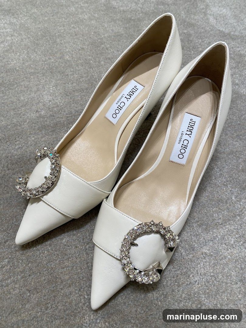 A pair of white leather pointed-toe pumps with crystal-embellished circular buckles on the vamp. The brand name 'JIMMY CHOO LONDON' is visible on the insoles.
