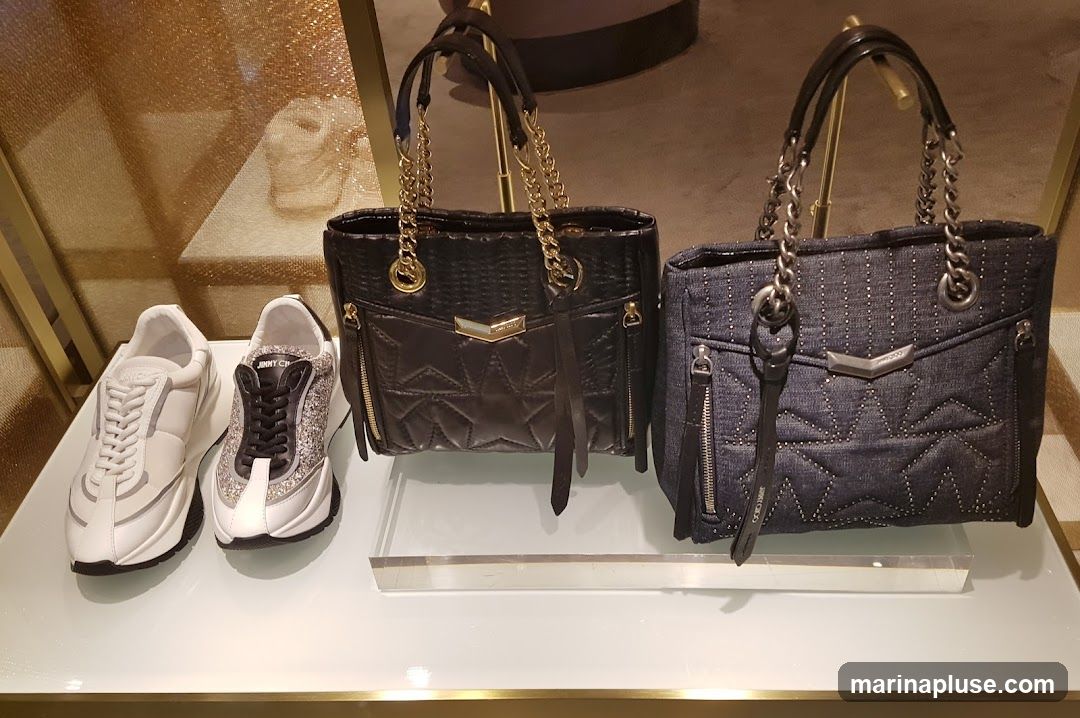 A display of two handbags and two pairs of sneakers on a white shelf. The handbags are black and denim, both with chain straps and decorative stitching. The sneakers are white and white with silver glitter accents. The brand name "JIMMY CHOO" is visible on the shoes and on a tag attached to the denim handbag.