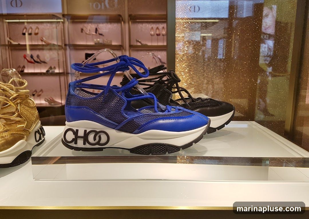 A close-up view of several pairs of designer sneakers displayed on a shelf in a retail store. In the foreground, a blue lace-up sneaker with a white sole and black logo detailing is prominently featured. To its left, a gold sneaker is partially visible. In the background, a black sneaker is also displayed. The store's interior is visible behind the shoes, with shelves holding more footwear and branding elements like the Dior logo.