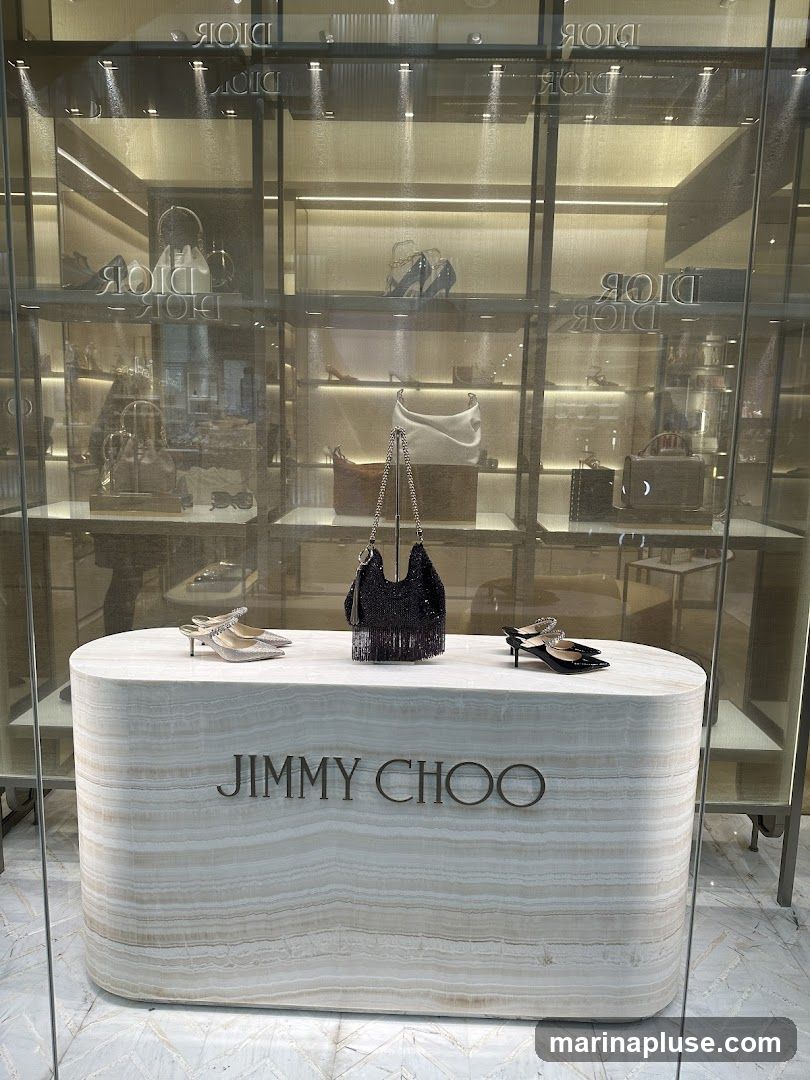 A storefront display for Jimmy Choo, featuring a marble counter with the brand name prominently displayed. On the counter, there are two pairs of women's shoes and a black beaded handbag. Behind the counter, shelves are stocked with various handbags and shoes, with the Dior logo visible on the glass in the background.