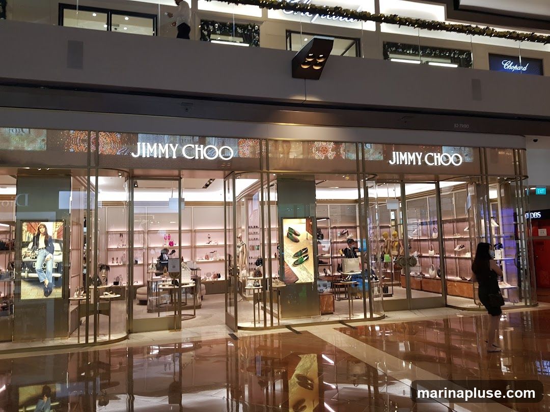 The exterior of a Jimmy Choo store with its name prominently displayed above the entrance. The store features large glass windows showcasing handbags and shoes on display shelves. A digital advertisement featuring a model wearing jeans and a jacket is visible on the left side of the entrance.
