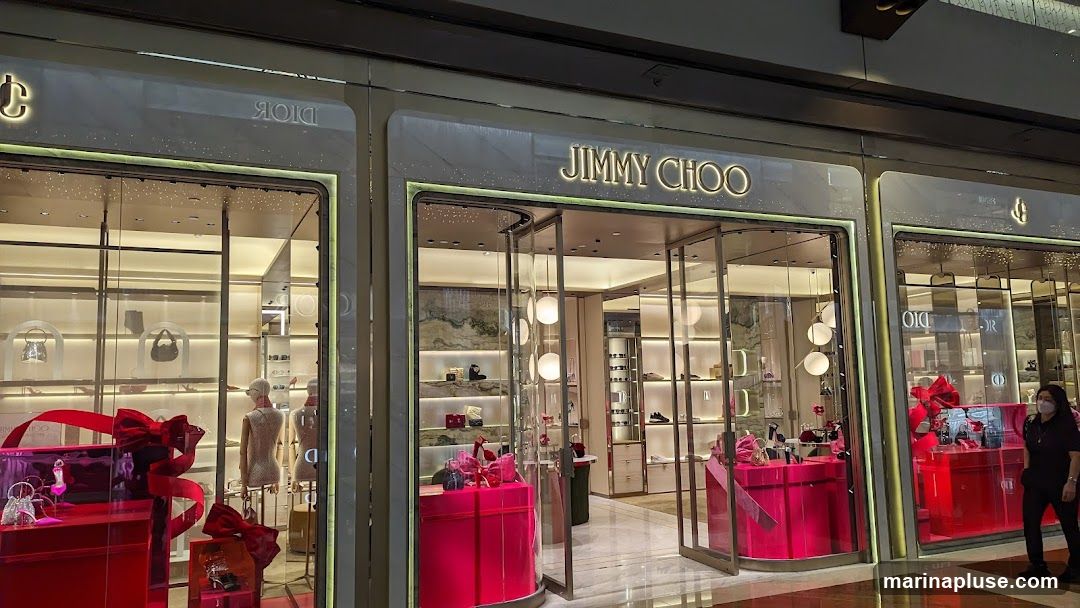 The exterior of a Jimmy Choo store. The storefront features large glass windows and doors, with the Jimmy Choo logo prominently displayed above the entrance. Inside, shelves display handbags and shoes, with mannequins and gift displays visible. The store is decorated with bright pink gift boxes and red ribbons.