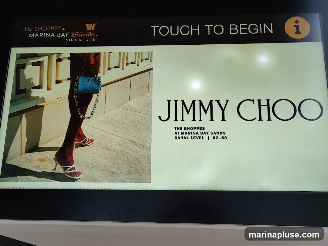 A digital display shows an advertisement for Jimmy Choo at The Shoppes at Marina Bay Sands, Singapore. The display features an image of a person's legs and feet wearing white sandals and holding a blue handbag. The Jimmy Choo logo is prominently displayed, along with the store's location. The text "TOUCH TO BEGIN" suggests it is an interactive kiosk.