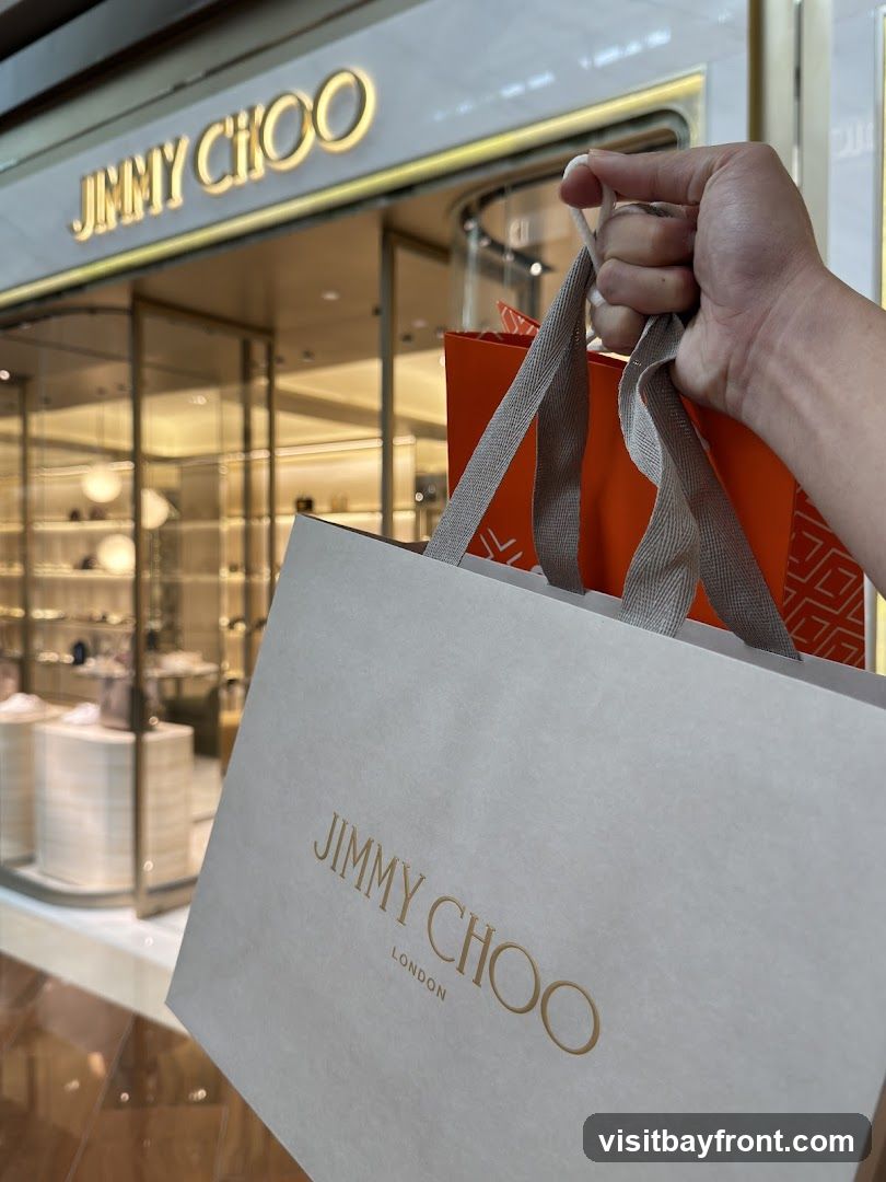 A hand holds shopping bags in front of a Jimmy Choo store. The store has a large, illuminated sign that reads "JIMMY CHOO" above a glass facade displaying merchandise. The shopping bags also feature the Jimmy Choo logo.