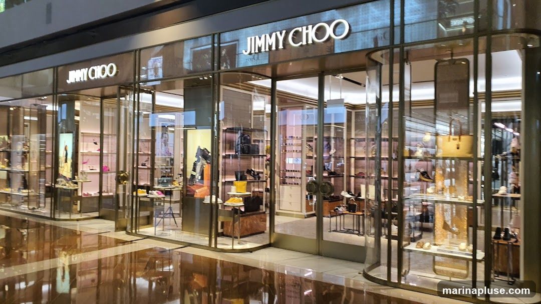 The exterior of a Jimmy Choo store, featuring a well-lit interior showcasing various handbags and shoes displayed on shelves and mannequins. The store has a modern design with glass panels and metallic accents. The prominent "JIMMY CHOO" logo is visible above the entrance.