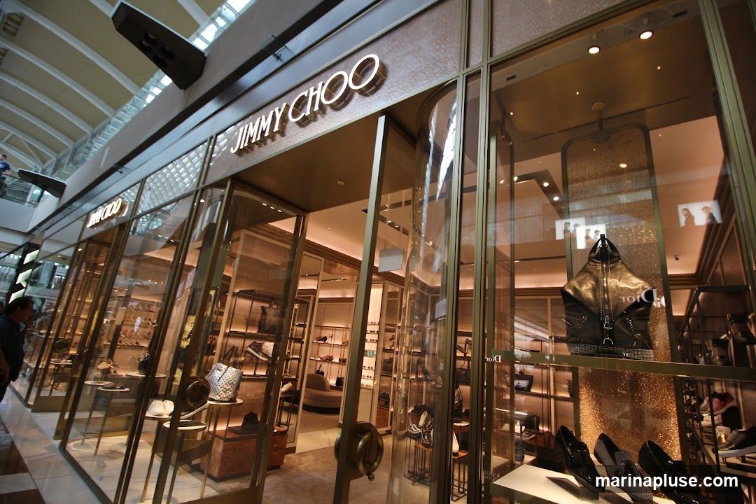 The exterior of a Jimmy Choo store with large glass windows displaying shoes and handbags. The store's name is prominently displayed in gold lettering above the entrance.