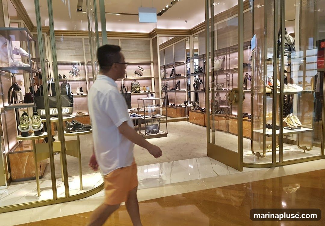 A man walks through a brightly lit, upscale shoe store. The store features glass display cases filled with various styles of shoes and handbags. Shelving units line the walls, showcasing more merchandise. The interior has a modern and luxurious design with marble flooring and gold accents.