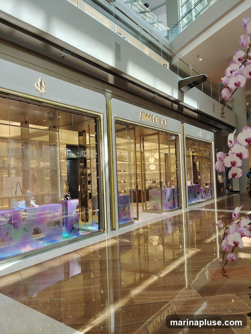 The exterior of a Jimmy Choo store in a modern shopping mall. The storefront features large glass windows displaying handbags and shoes. The store's name, JIMMY CHOO, is prominently displayed in gold lettering above the entrance. The interior of the store is brightly lit, showcasing merchandise on shelves. A decorative display with iridescent panels is visible in the window. To the right, a branch of pink and white orchids adds a touch of nature to the scene. The polished floor reflects the light and the store's facade.