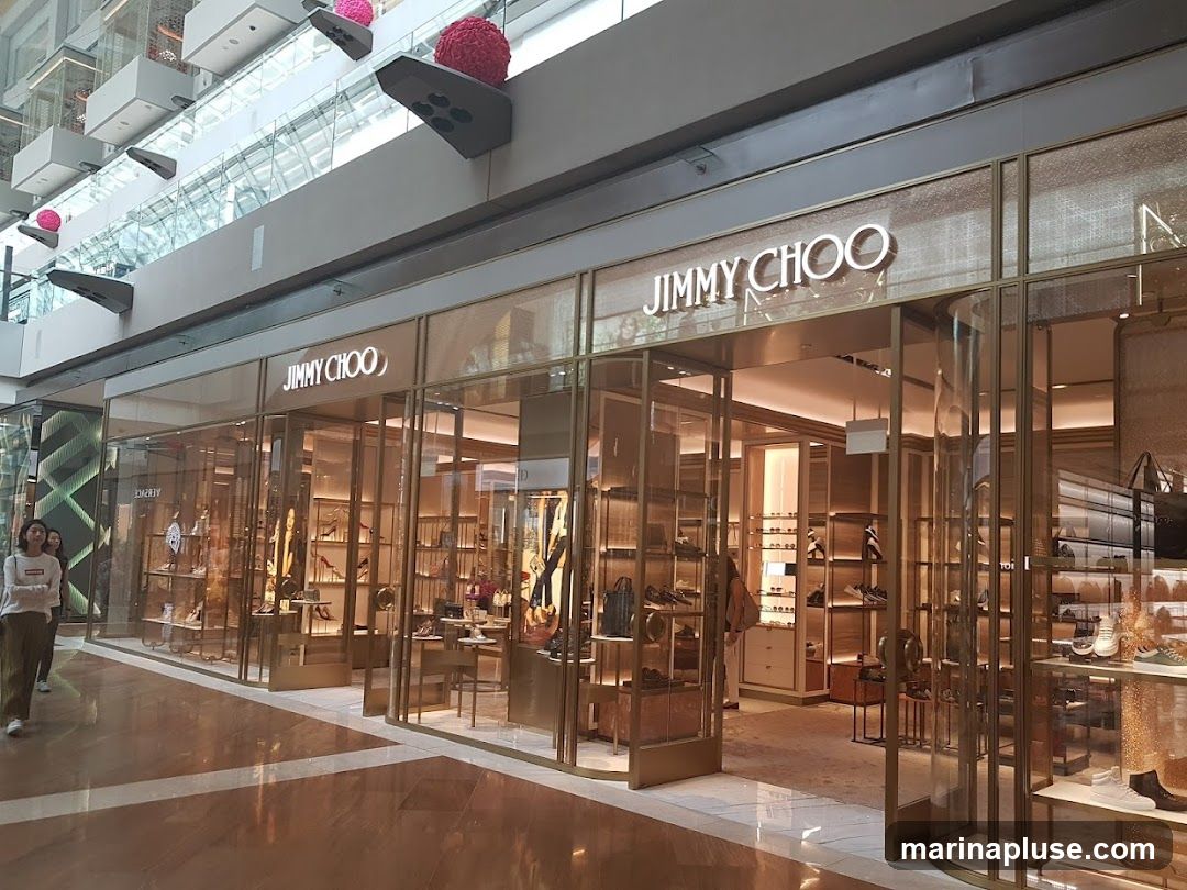 A well-lit Jimmy Choo store with a polished facade. The store displays a variety of shoes and handbags on shelves behind glass windows. The brand name "JIMMY CHOO" is prominently displayed in large, illuminated letters above the entrance. Two women are walking past the storefront on the left.