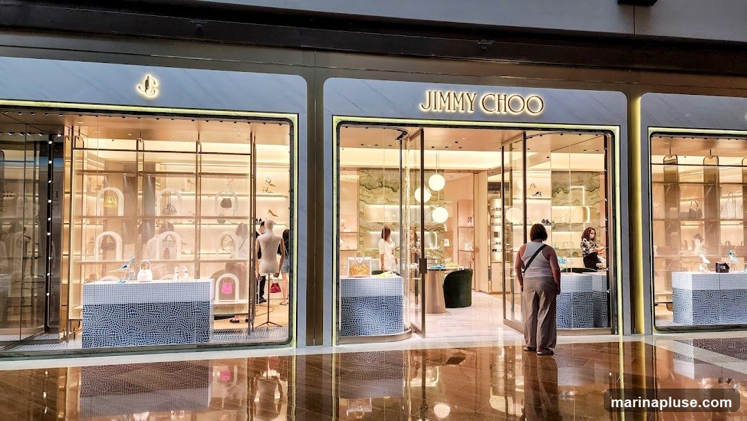 The exterior of a Jimmy Choo store with its name prominently displayed above the entrance. The store features large glass windows showcasing handbags and shoes displayed on shelves and mannequins. The interior is brightly lit with a modern and luxurious aesthetic. Several customers and staff members are visible inside.