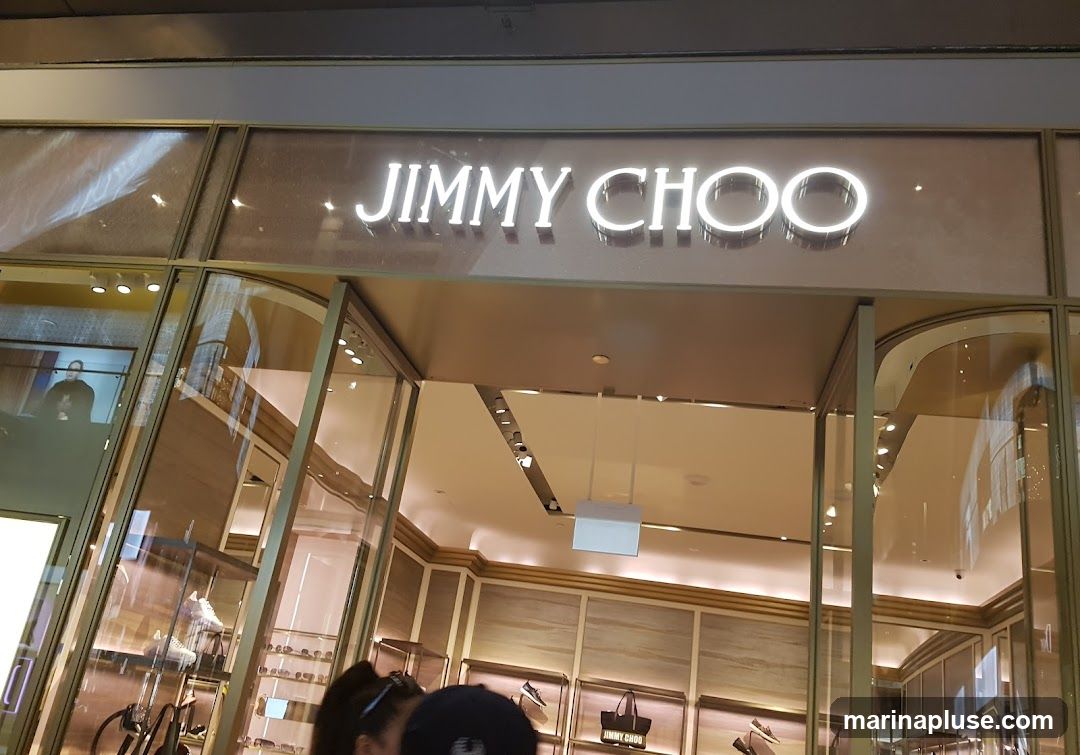 The exterior of a Jimmy Choo store with the brand name illuminated above the entrance. The store features large glass windows displaying merchandise, including shoes and handbags. The interior is visible through the windows, showing shelves and lighting.