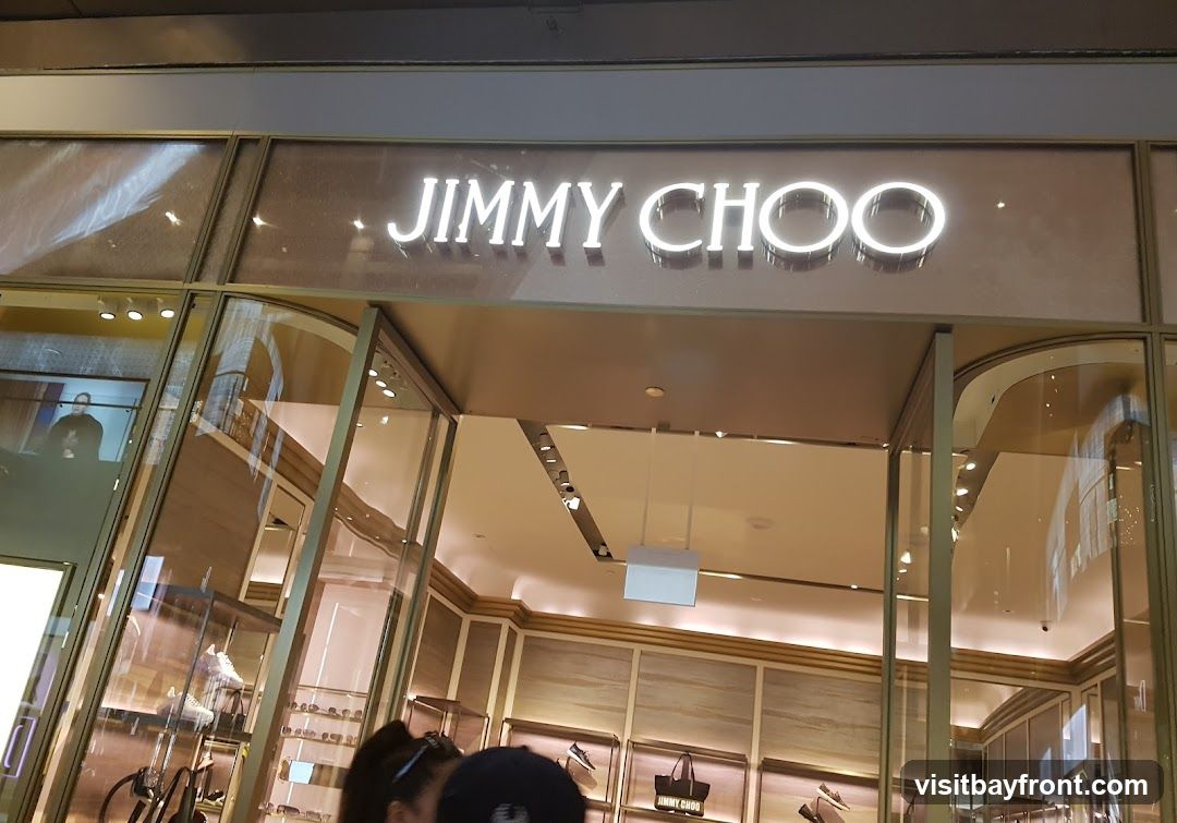 The exterior of a Jimmy Choo store with the brand name illuminated above the entrance. The store features large glass windows displaying merchandise, including shoes and handbags. The interior is visible through the windows, showing shelves and lighting.