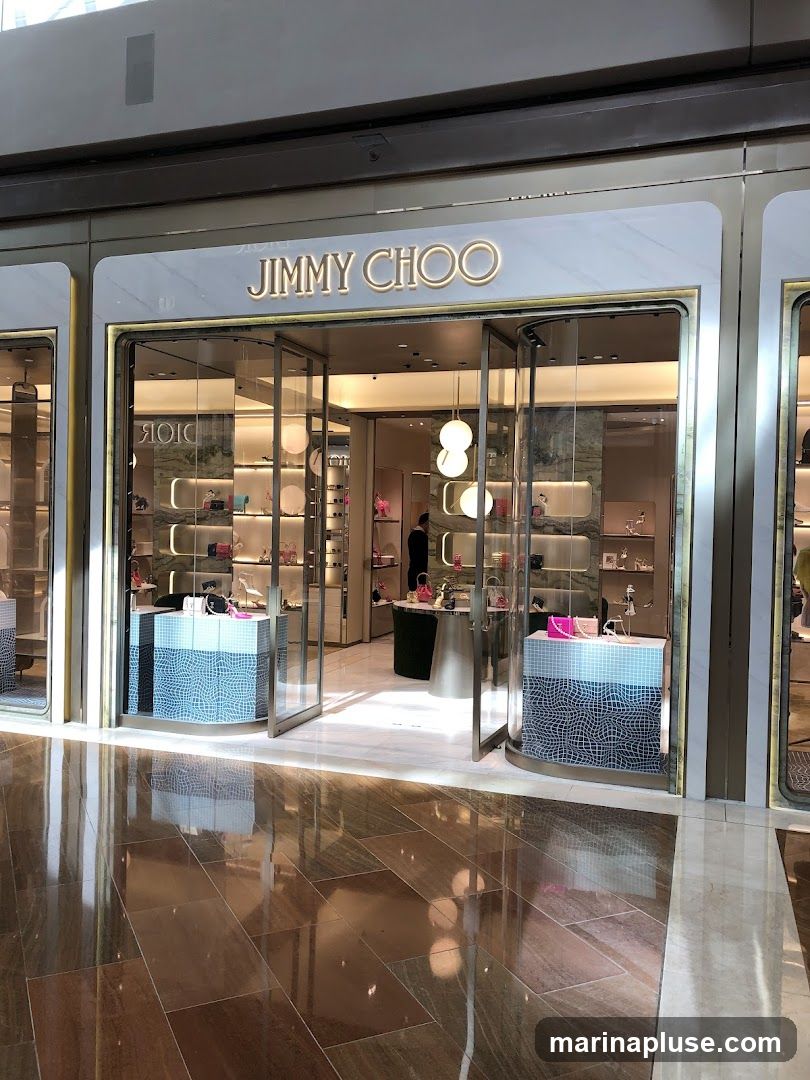 The exterior of a Jimmy Choo store with its name prominently displayed above the entrance. The store features a modern design with glass doors and windows showcasing handbags and shoes on display shelves. The floor in front of the store is highly reflective.