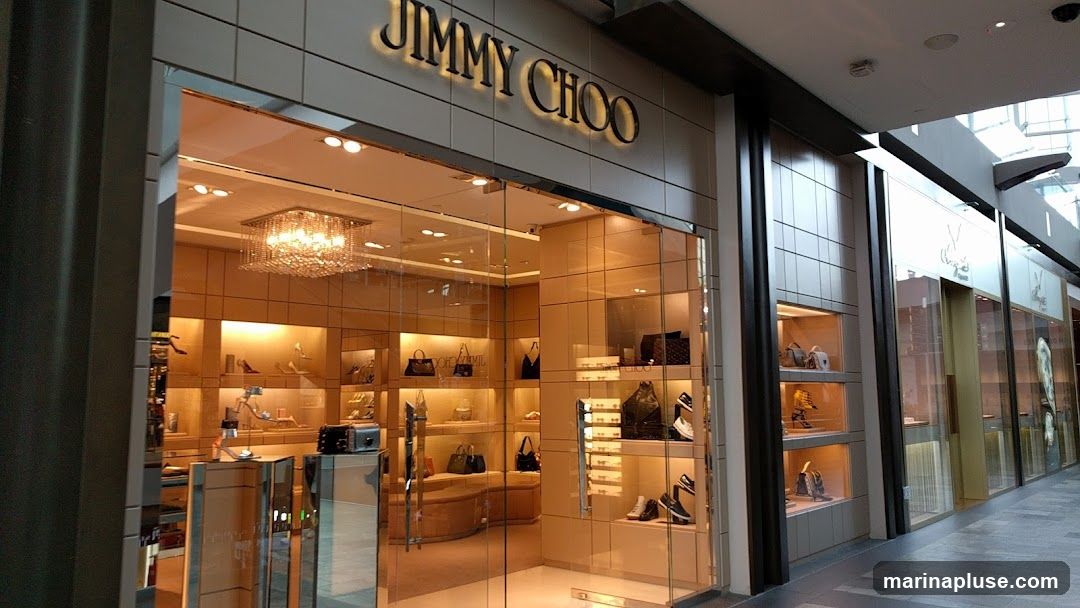 A well-lit luxury store with a prominent "JIMMY CHOO" sign above a glass facade. The interior displays shoes and handbags on illuminated shelves. The store appears to be located in a modern shopping mall with a tiled floor and a skylight.