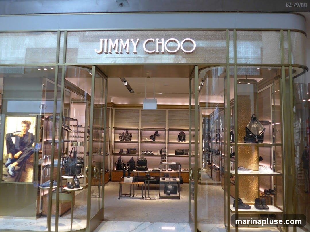 The exterior of a Jimmy Choo store with large glass windows and doors. The store's name is prominently displayed in illuminated letters above the entrance. Inside, shelves display various handbags and shoes. A large advertisement featuring a man is visible on the left side.