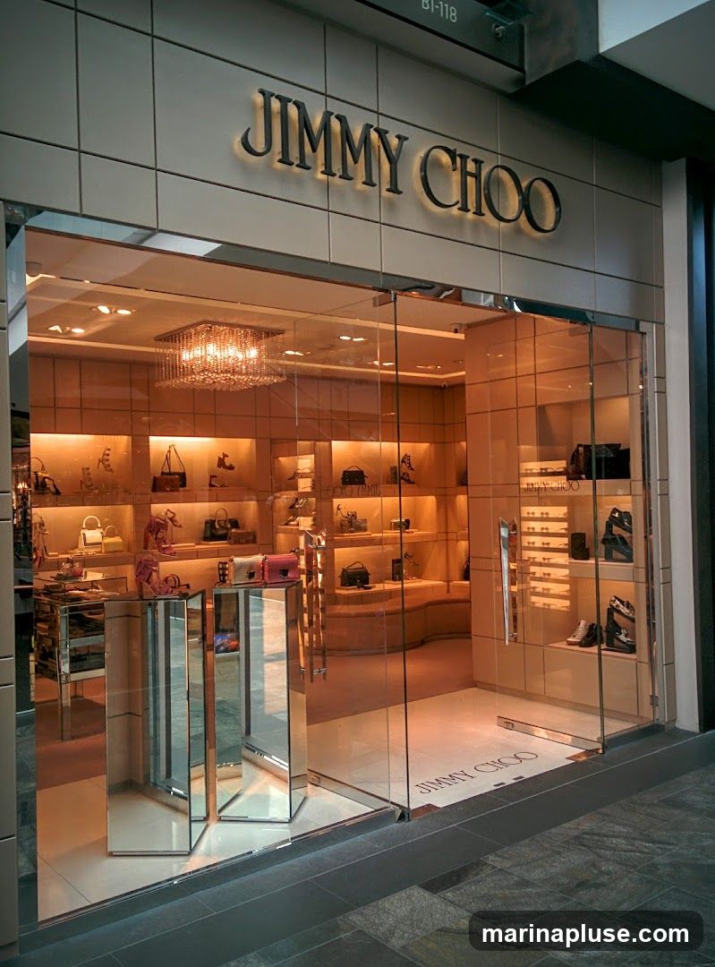 The exterior of a Jimmy Choo store with a prominent "JIMMY CHOO" sign above the entrance. The store features a modern glass facade with a large display window showcasing handbags and shoes. Inside, shelves are illuminated, displaying various luxury accessories.