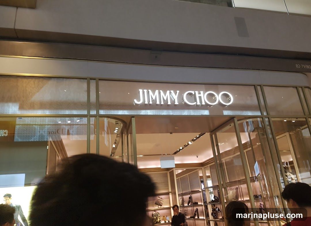 The exterior of a Jimmy Choo store with the brand name illuminated above the entrance. The store features a modern design with glass windows displaying shoes and accessories inside.