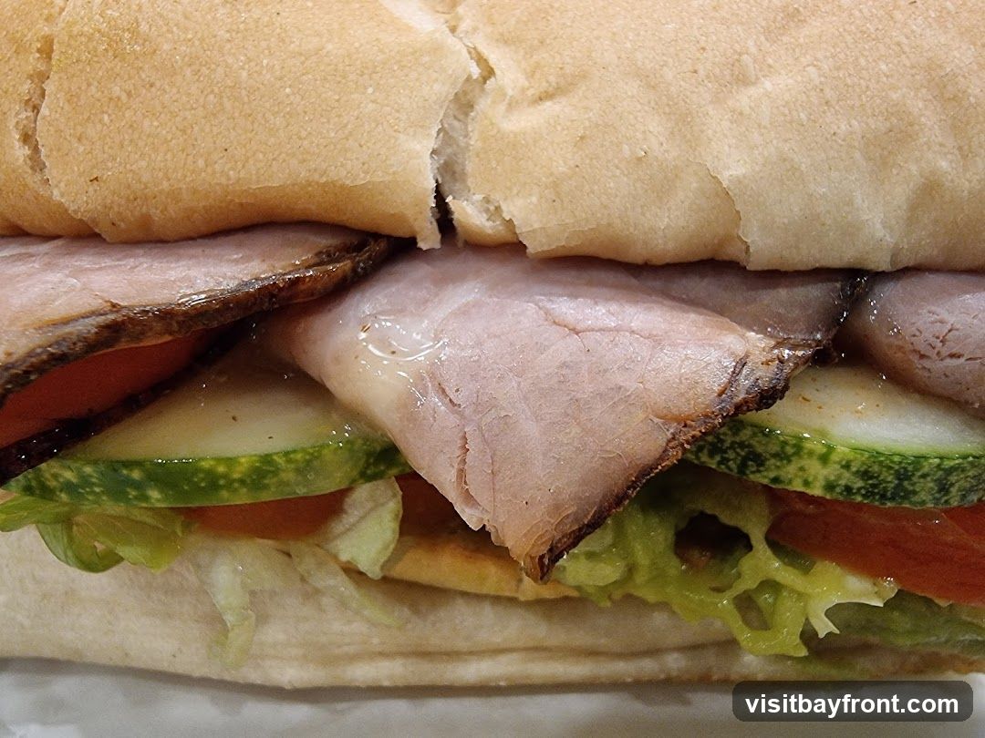 A close-up, detailed view of a sandwich. The sandwich is made with a long, soft bun, sliced thinly. Inside, layers of thinly sliced roast meat, red tomato slices, crisp green lettuce, and round cucumber slices are visible. The meat appears to be well-cooked with some browned edges.