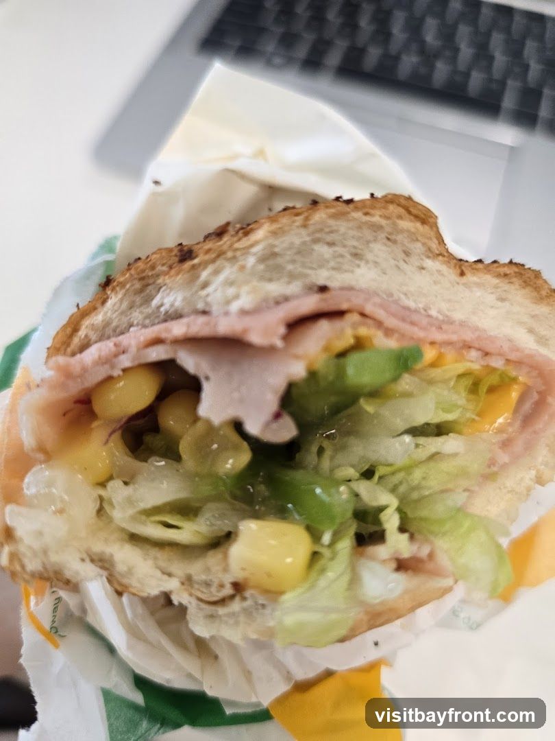 A close-up shot of a sandwich with ham, cheese, corn, and lettuce, wrapped in paper. The sandwich is on a toasted bun. A laptop keyboard is visible in the blurred background.
