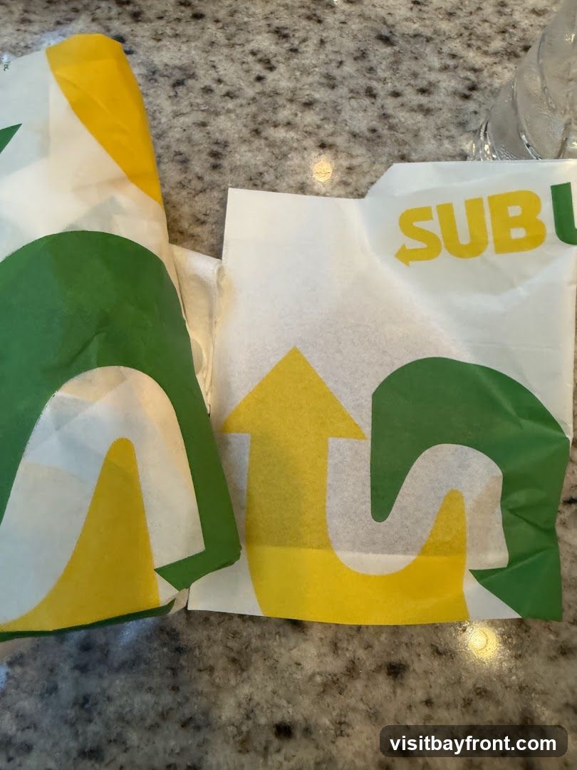 Two food wrappers with the Subway logo and branding are laid out on a speckled countertop. The wrappers are white with green and yellow accents, featuring the distinctive Subway arrow design.