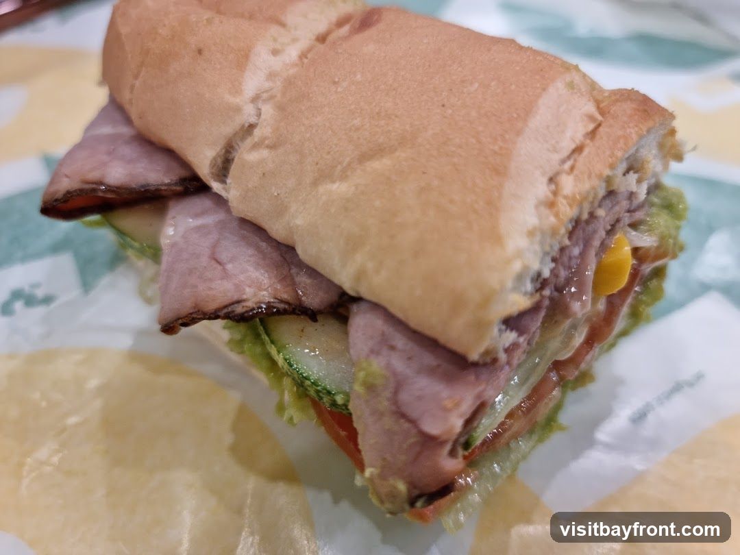 A close-up shot of a roast beef sub sandwich on a white paper wrapper with a green and white pattern. The sandwich is made with a long, light-colored bun, filled with slices of roast beef, lettuce, tomato, cucumber, and what appears to be guacamole and corn. The sandwich is cut in half, revealing the layers of ingredients.